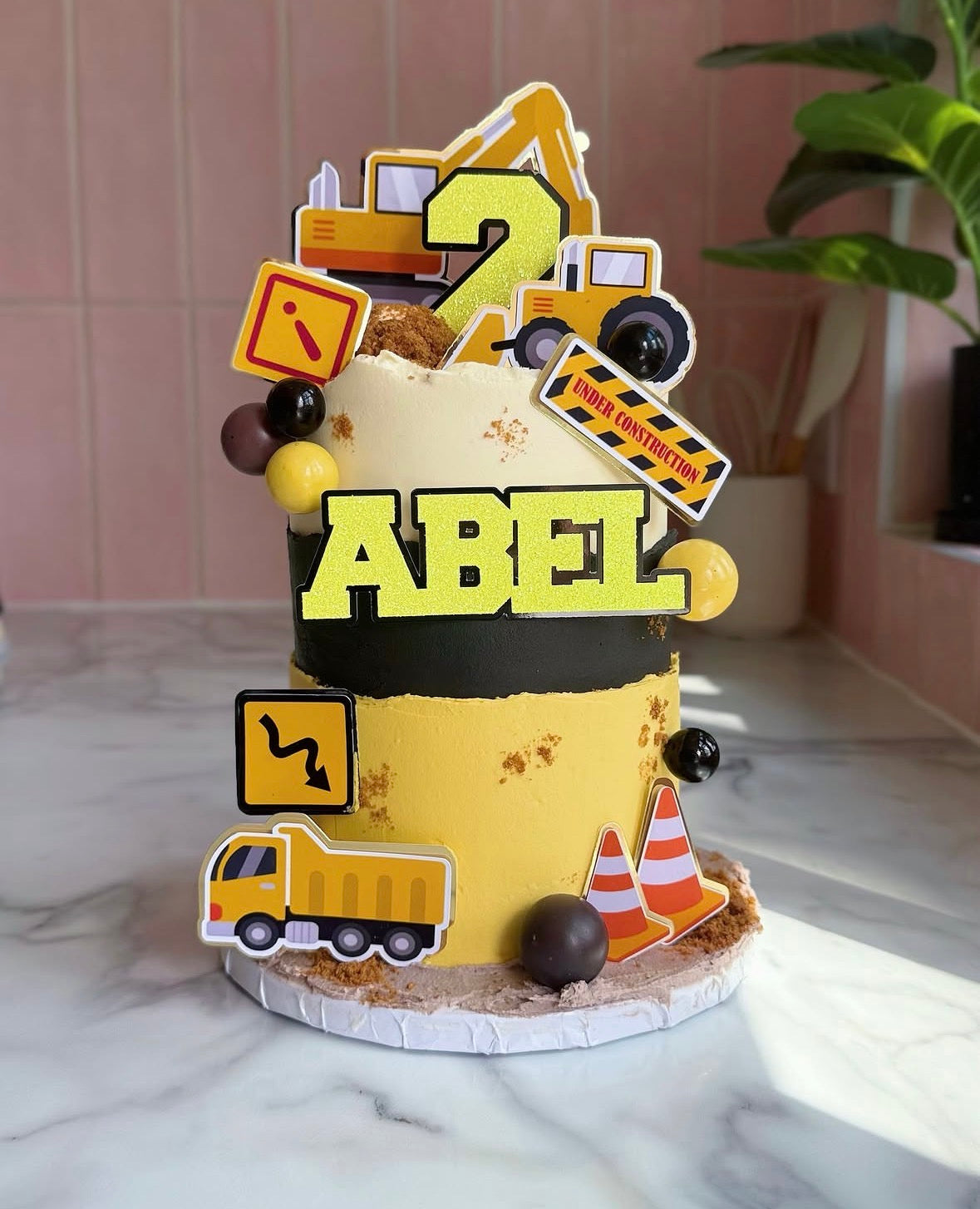 A custom construction-themed birthday cake decorated with yellow and black frosting, a large 'ABEL' name topper, and various toy construction vehicle and sign toppers, displayed on a marble countertop.
