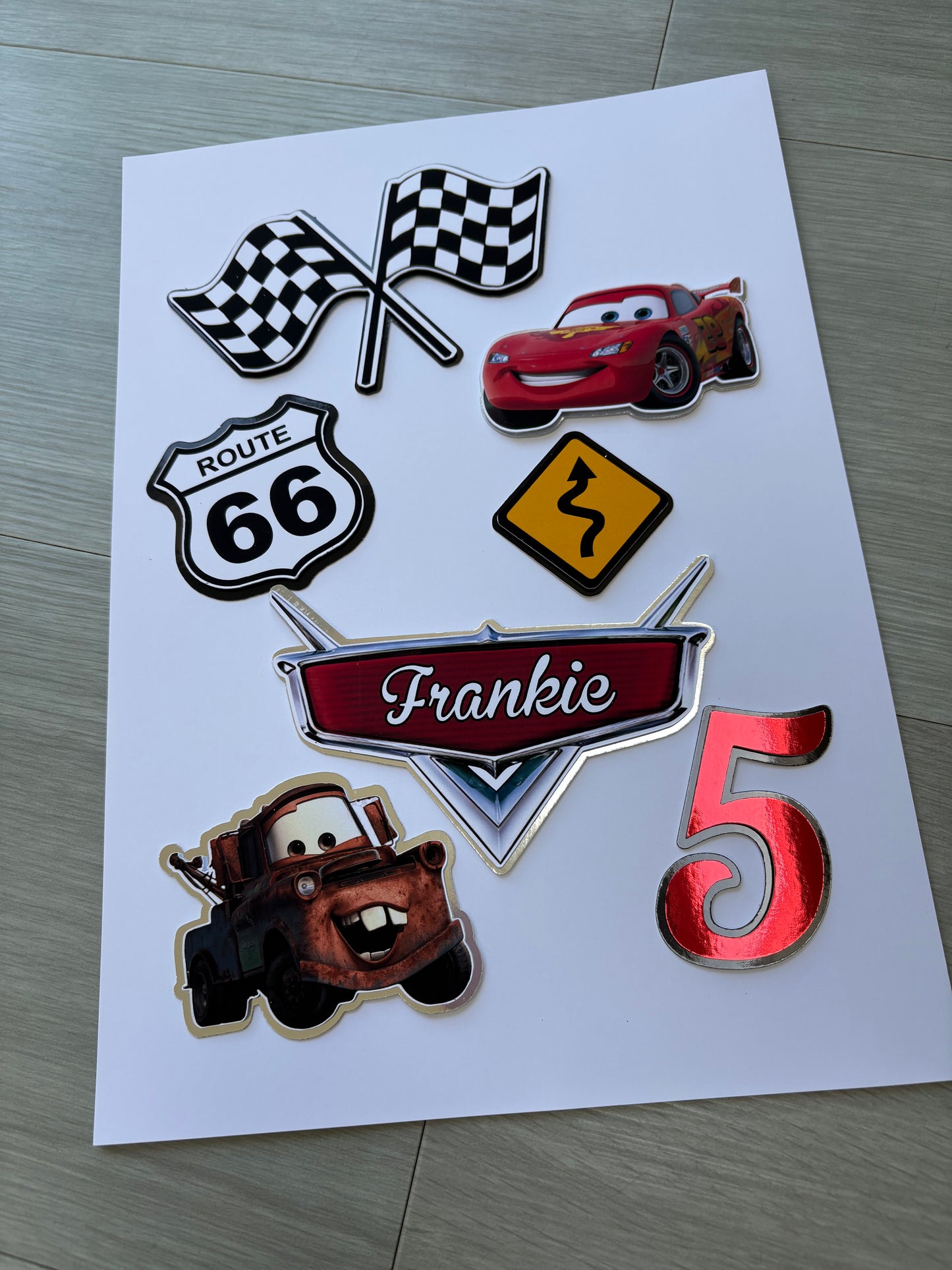 A sheet of custom metallic die-cut stickers featuring characters and motifs from the Cars franchise, including Lightning McQueen, Mater, a checkered flag, a Route 66 shield, a speed limit sign, and a red number 5, displayed on a light gray wood floor.