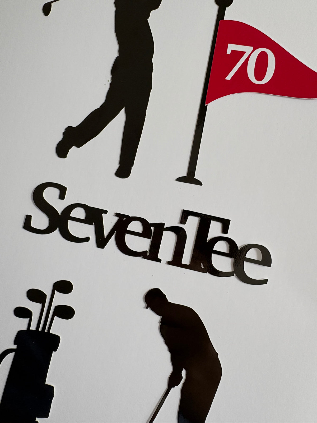 A close-up of a golf-themed cake topper set wall  featuring black silhouettes of golfers, a golf bag, and a flag with the number 70, above the word 'SevenTee' in black lettering on a white background.