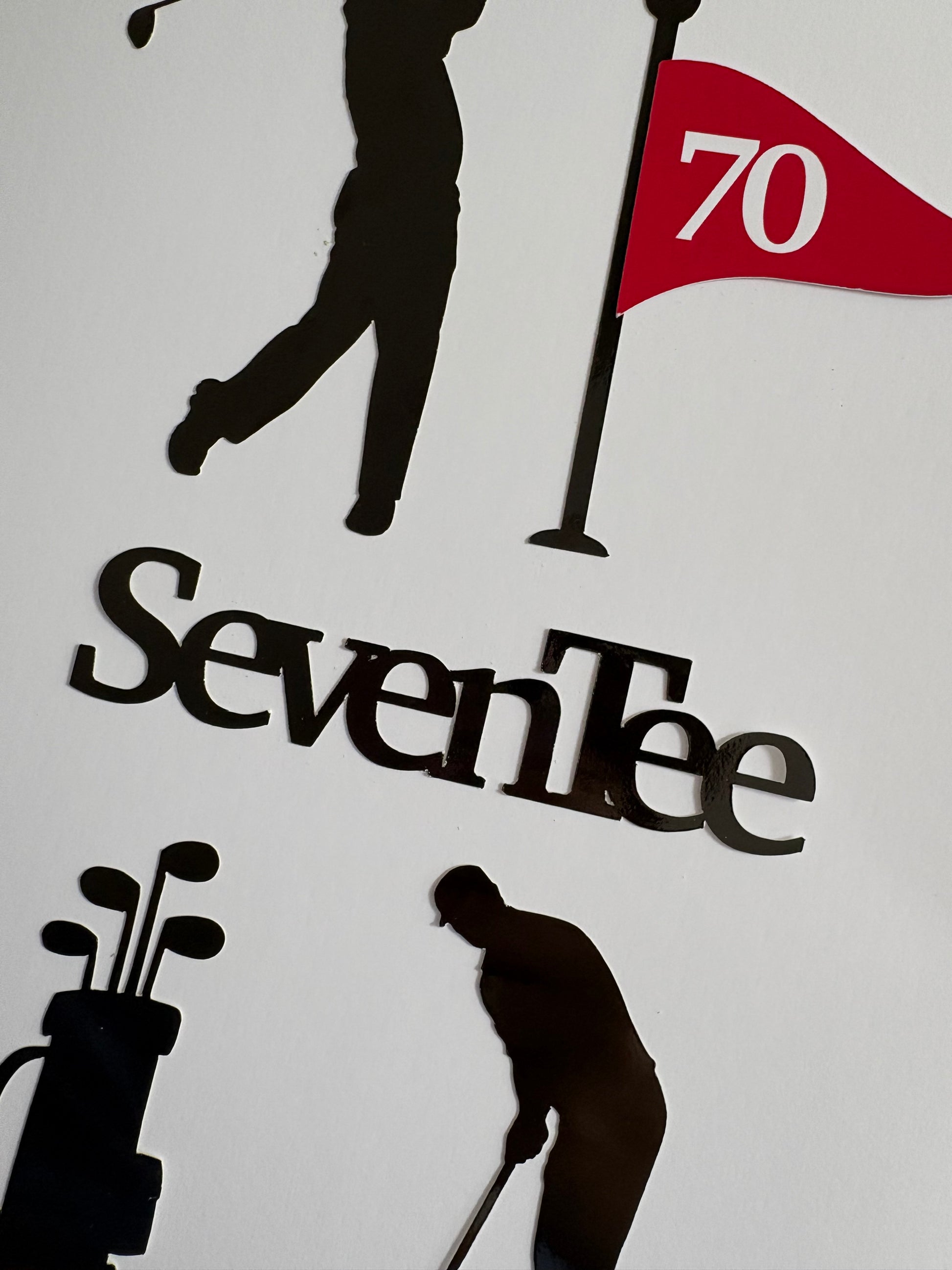A close-up of a golf-themed cake topper set wall  featuring black silhouettes of golfers, a golf bag, and a flag with the number 70, above the word 'SevenTee' in black lettering on a white background.