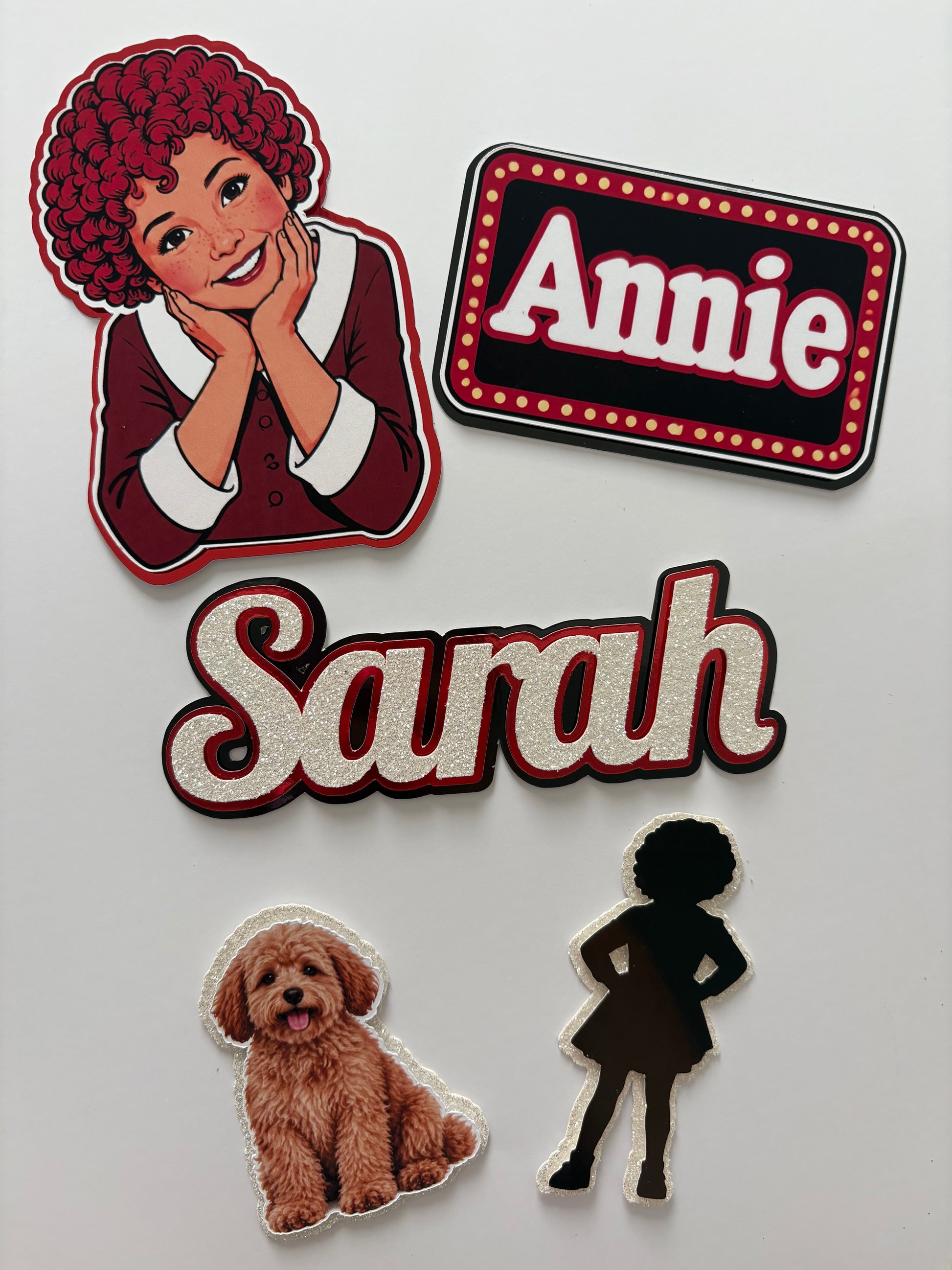 A set of personalised cake toppers on a white background, featuring a portrait of a woman with curly red hair labeled 'Annie', a large glittery 'Sarah', a brown dog illustration, and a black silhouette patch.