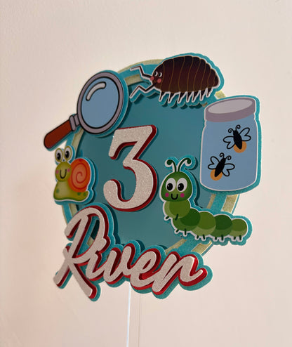 A decorative personalized birthday sign featuring the name '3 River' with insect illustrations including a magnifying glass, beetle, snail, caterpillar, and jar of flies, mounted on a stick against a plain wall.