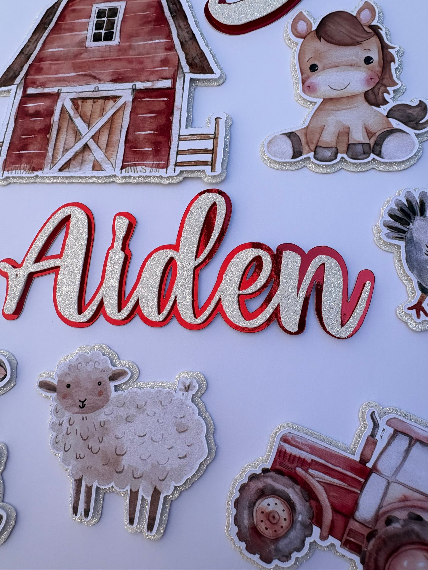 Personalized farm-themed paper cutout decorations featuring the name 'Alden' in glittery white script with a red outline, surrounded by illustrations of a barn, sheep, tractor, and a cartoon dog on a white background.