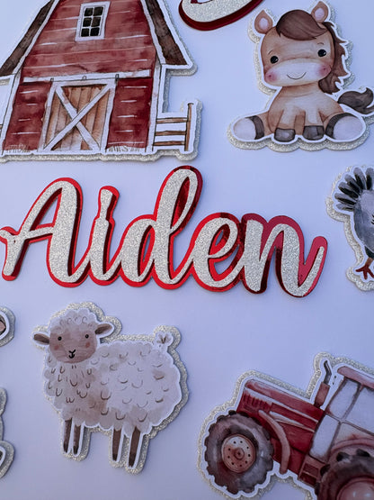 Personalized farm-themed paper cutout decorations featuring the name 'Alden' in glittery white script with a red outline, surrounded by illustrations of a barn, sheep, tractor, and a cartoon dog on a white background.