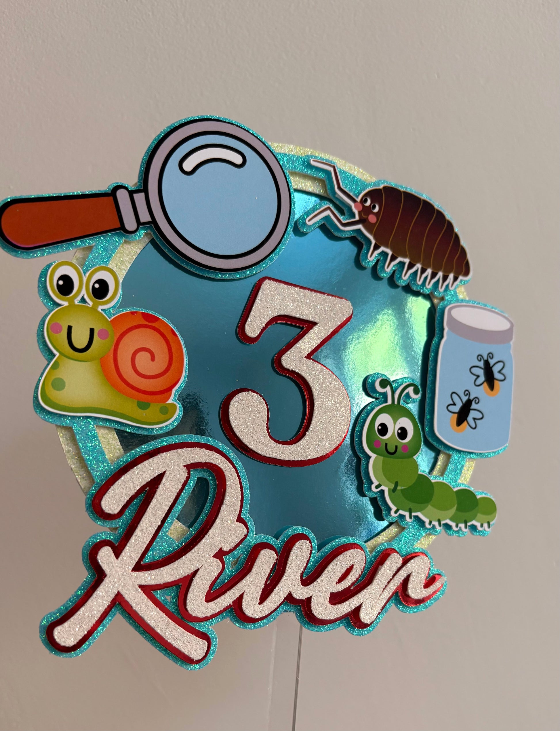 A decorative personalized sign reading '3 River' with a blue glittery background and cartoon-style bugs and nature illustrations including a magnifying glass, snail, beetle, caterpillar, and bug jar.