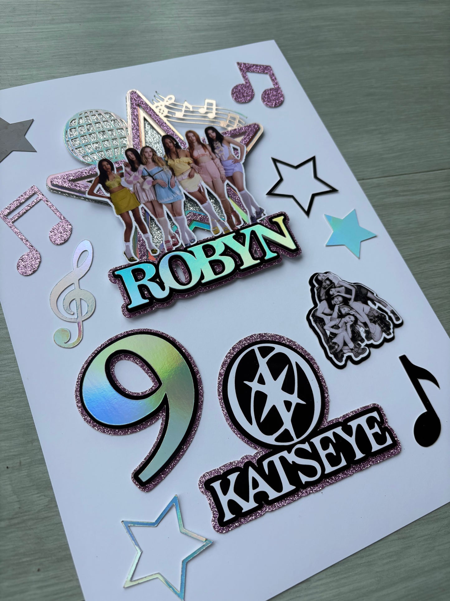 A sheet of decorative cake toppers or embellishments on a white backing card, featuring musical notes, stars, a treble clef, a large holographic number 9, and text stickers reading 'NABOR' and 'KATSEYE’ on a light wood surface.
