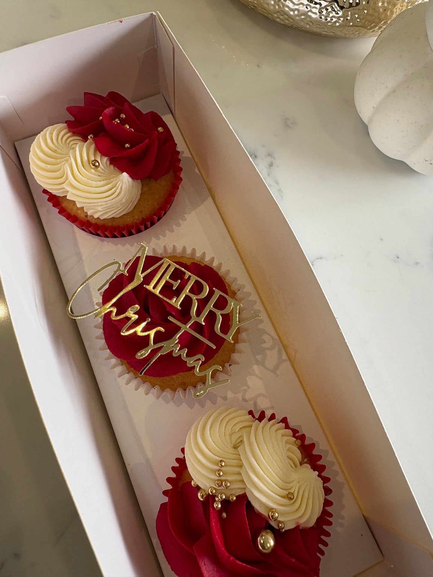 Three decorated holiday cupcakes in a white bakery box on a marble countertop. Two cupcakes have cream swirl frosting topped with red roses, and the center cupcake has red frosting with a gold 'Merry Christmas' topper.