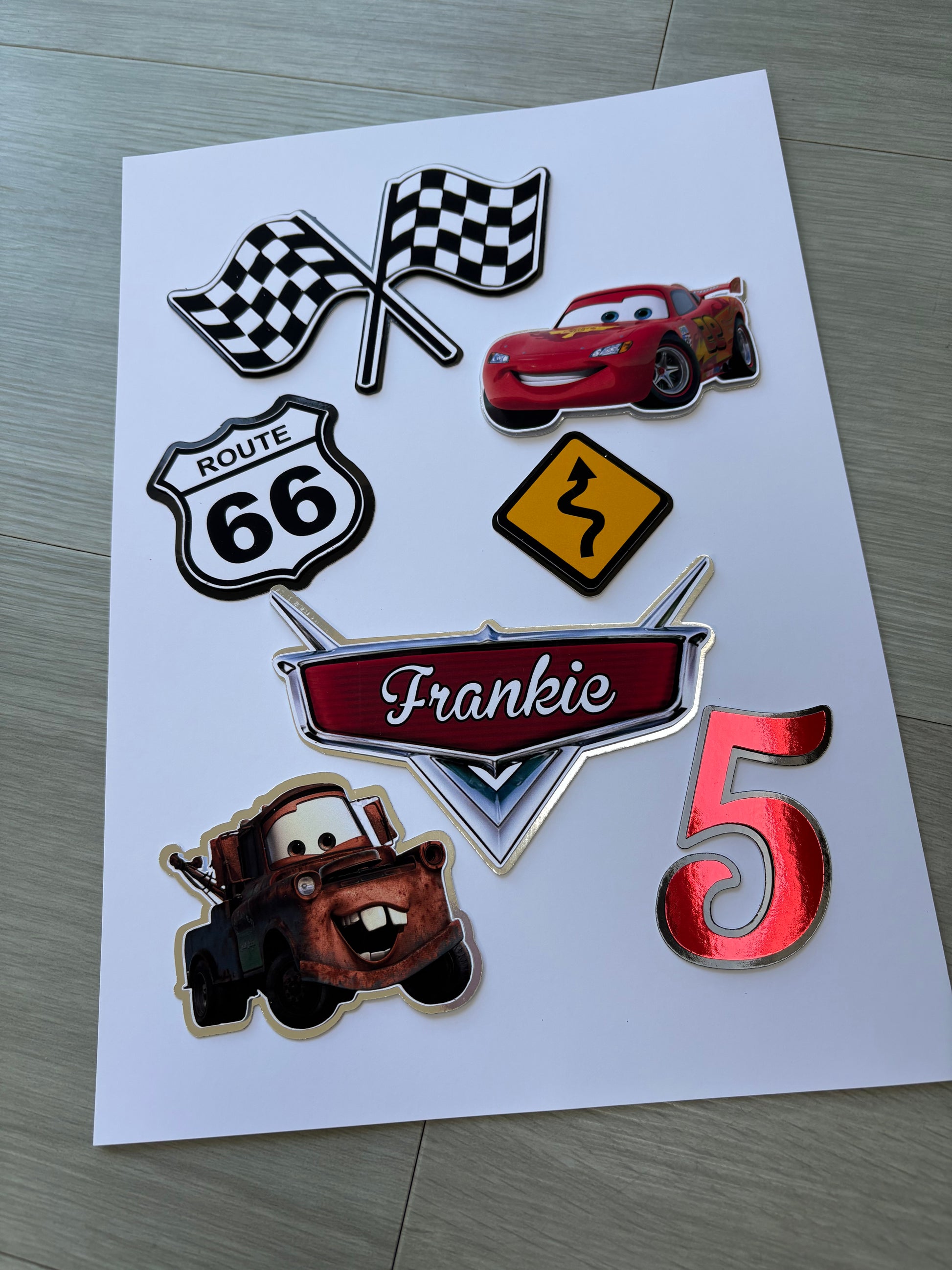 A sheet of custom metallic die-cut stickers featuring characters and motifs from the Cars franchise, including Lightning McQueen, Mater, a checkered flag, a Route 66 shield, a speed limit sign, and a red number 5, displayed on a light gray wood floor.