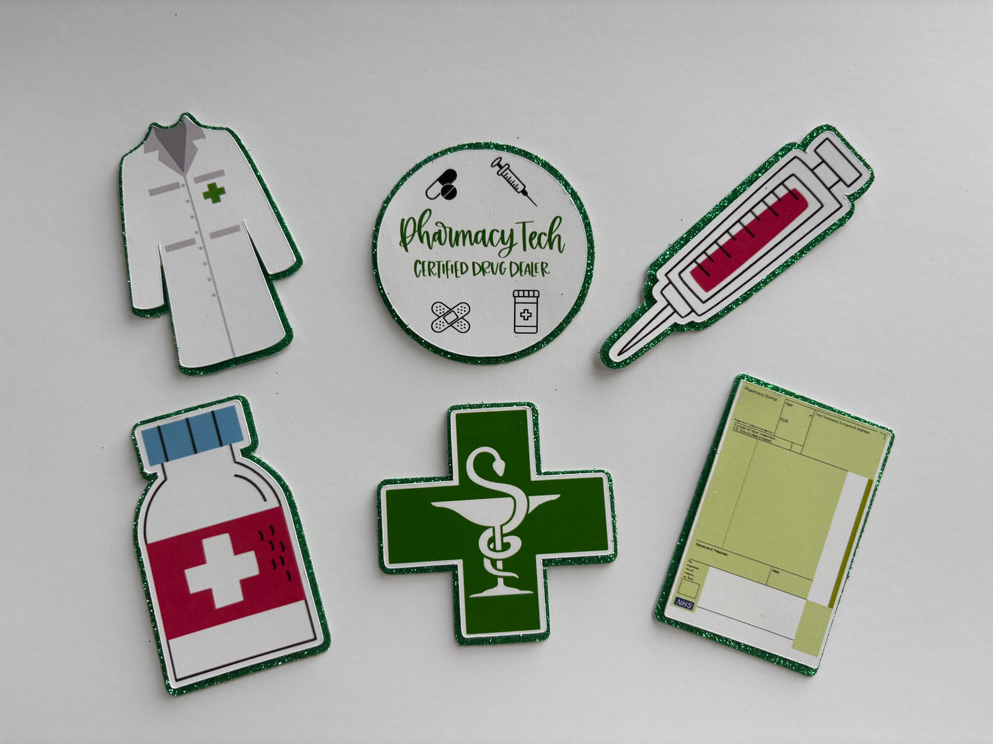 Six die-cut paper craft templates or stickers featuring pharmacy and medical designs, including a white lab coat, a circular 'Pharmacy Tech' badge, a syringe, a medicine bottle, a green cross with a snake and staff of Asclepius, and a prescription pad, arranged on a light gray background.