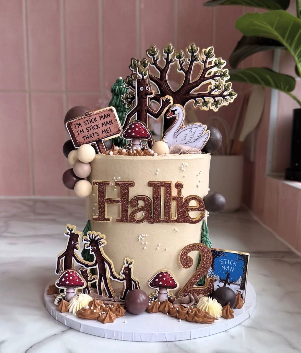 A decorated birthday cake with woodland-themed toppers, including a tree, swan, mushrooms, reindeer, and sign reading 'Hattie 2'.