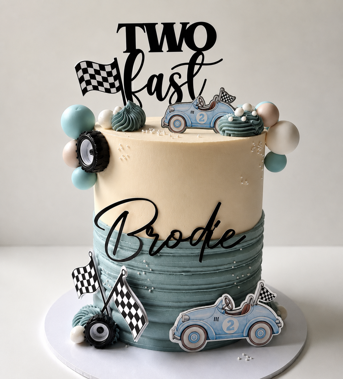 A custom two-year birthday cake decorated with racing-themed elements. The cake has a cream-colored top half and a textured light blue-green bottom half.