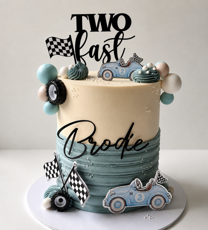 A custom two-year birthday cake decorated with racing-themed elements. The cake has a cream-colored top half and a textured light blue-green bottom half.