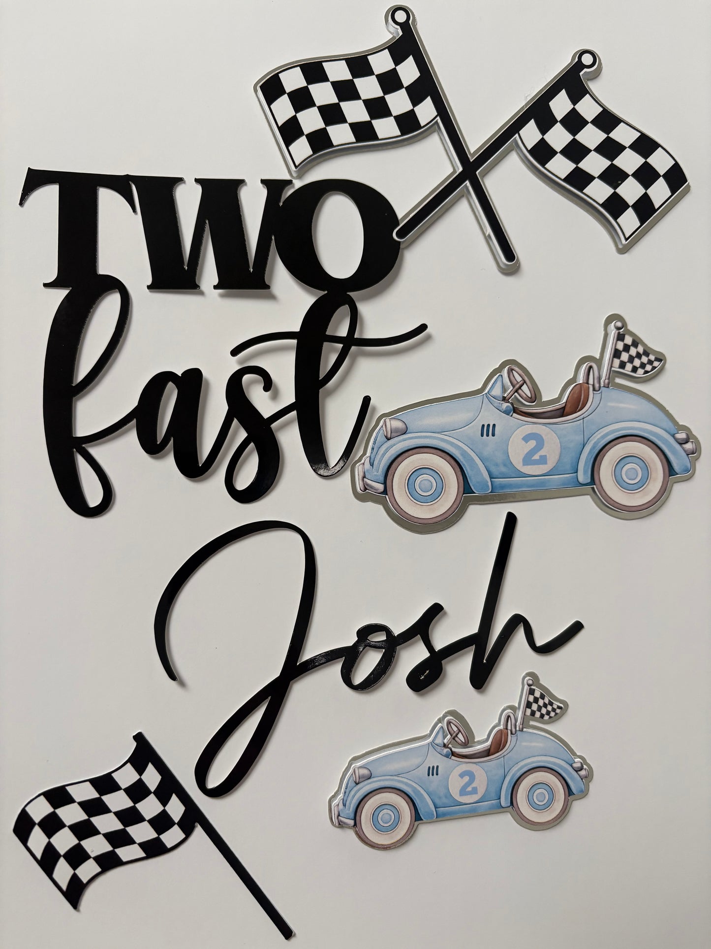 A set of custom racing-themed cake toppers on a light gray background, featuring two blue race cars, checkered flags, and black script lettering that reads 'Two Fast Josh'.