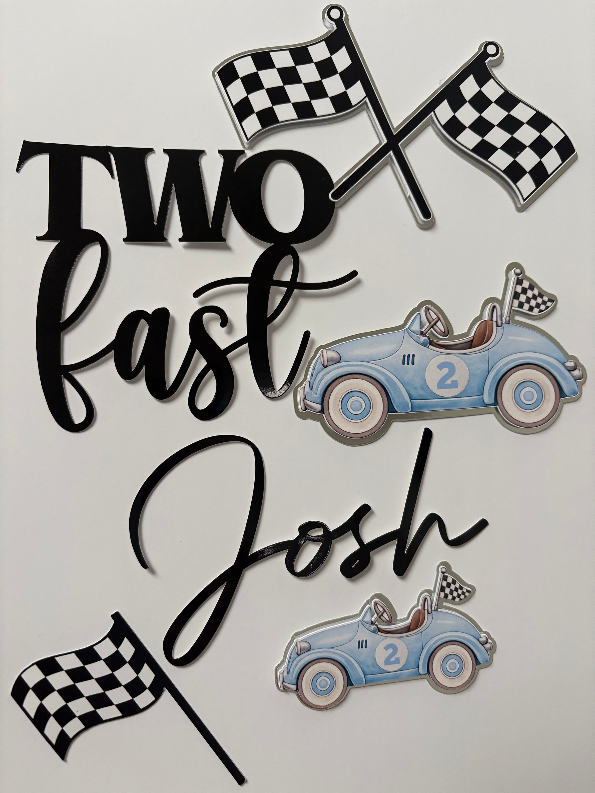 A set of custom racing-themed cake toppers on a light gray background, featuring two blue race cars, checkered flags, and black script lettering that reads 'Two Fast Josh'.