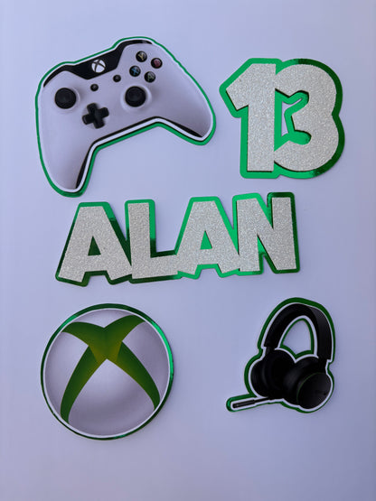 A set of Xbox-themed party decorations or cutouts on a light lavender background, including a white and black game controller, a green and white number 13, the name 'ALAN' in glittery green letters, a black headset, and a round patch with the green Xbox logo.